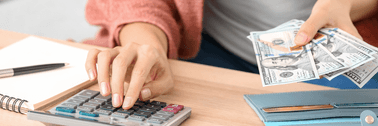 A woman in a pink cardigan uses a calculator on her desk while holding a stack of cash in front of her wallet.