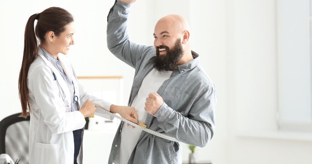 happy female doctor using tape to measure waist of male client at clinic