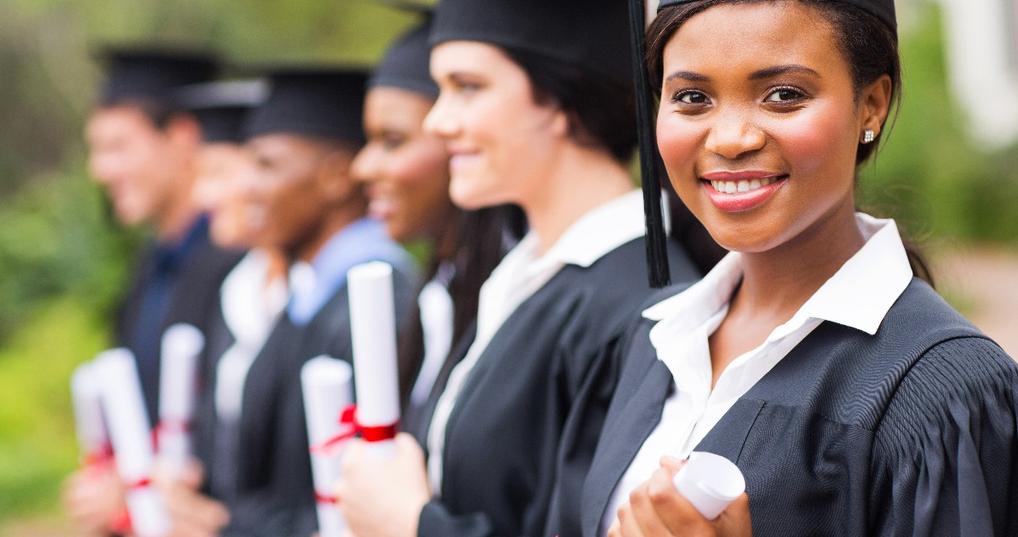 african american female student wearing graduation gown and hat with other students in background at graduation ceremony in college