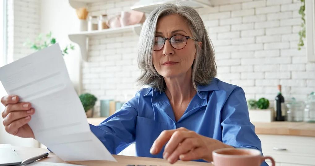 senior mature business woman holding paper bill using calculator managing account finance while sitting at home kitchen table