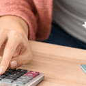 A woman in a pink cardigan uses a calculator on her desk while holding a stack of cash in front of her wallet.