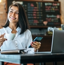 Woman working in a coffee shop. She's holding a phone in one hand and a cup of coffee in the other. 