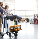 Woman sitting on luggage on a cart being pushed by a man through an airport