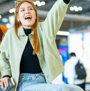 A woman rides atop a luggage cart in the airport while her friend pushes it from behind.