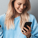 A woman in a blue sweater smiles while holding her phone and credit card. 