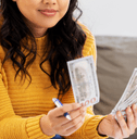 A woman in a yellow sweater sits on the couch while counting a stack of 100 dollar bills.