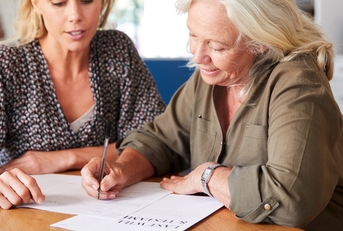 Female Friend Helping Senior Woman To Complete Last Will And Testament At Home