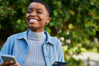 A smiling woman holds a credit card in one hand and a smartphone in the other. 