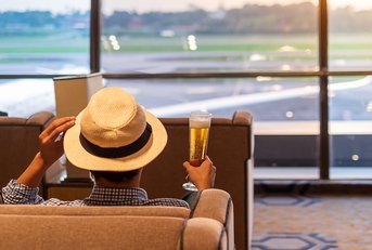 A man sits in a chair at the airport holding a beer and touching the brim of his hat. 