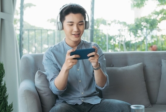 A man wearing headphones sits on a couch and plays mobile games on his phone. 