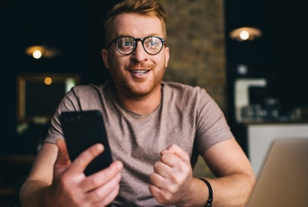 A man wearing glasses sits in front of a laptop and holding a smartphone. 
