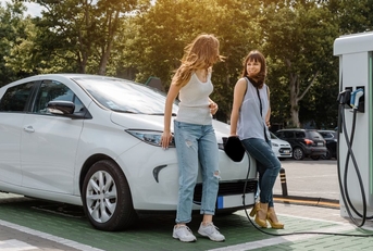Two girls leaning on white electric car waiting while it's charging at station green trees and sun 