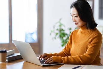 asian woman happily using laptop on table with coffee and files at workspace