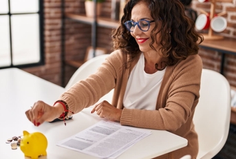 middle age woman sitting at table with papers putting pennie into yellow piggy bank