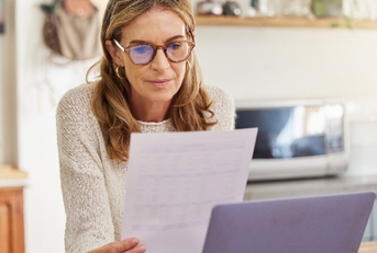 woman sitting at counter top in kitchen reviewing documents while using computer at home