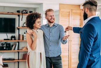 young male real estate agent standing standing inside house handing keys to couple