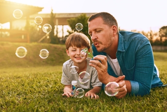 father and son leaning on lawn outdoors playing together while blowing bubbles