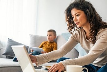 A mom works from her laptop while sitting on the couch next to a young child. 