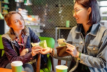 asian woman lending money to girl sitting inside cafe 