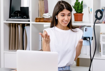 A content creator sits at a desk facing a phone held on a tripod. She is making a video to post on social media. 