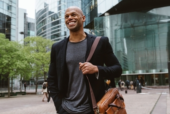 happy african american man walking to the office building, while smiling and carrying a messenger bag.
