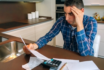 man sitting in home kitchen holding head while stressing about bills at countertop