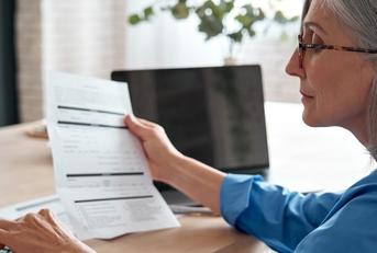 senior lady holding paper bill and calculating the monthly expenses on the calculater while sitting at the table at home.