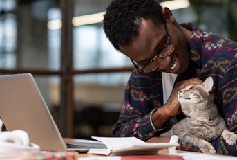 A young black man working on a laptop while holding his cute grey cat in his hands.