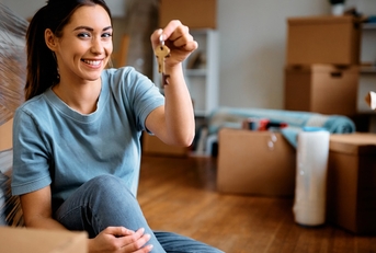 a young woman, brimming with joy, holding a key while moving into her new home with cardboard boxes filled with packed luggage.