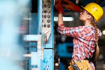 Female technician wearing yellow safety halmet is fixing a part of machinery with screwdriver in factory.