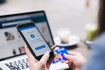 A woman is holding a smartphone with a screen that says payment success in the foreground. In the background, you can see a laptop and a credit card.