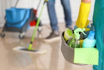 Female janitor holding a basket of cleaning supplies in the foreground with another janitor mopping in the background.l