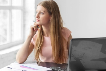 A young adult woman sits and thinks at a desk with a laptop.