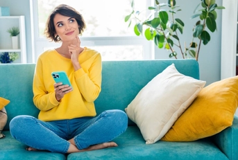 A girl is thinking while sitting on the couch in the living room and holding her phone.