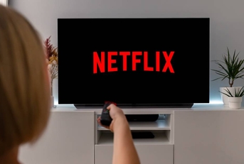 A woman holds up her remote control pointing to her TV, which is showing the Netflix screen.