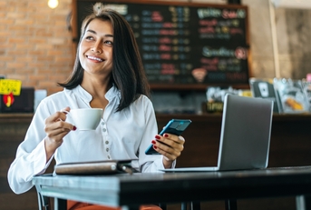 Woman working in a coffee shop. She's holding a phone in one hand and a cup of coffee in the other. 