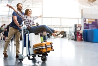 Woman sitting on luggage on a cart being pushed by a man through an airport