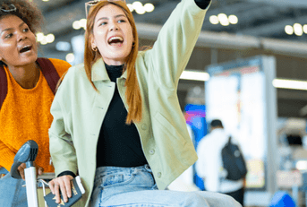 A woman rides atop a luggage cart in the airport while her friend pushes it from behind.