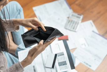 A woman opens an empty black wallet, while bills and paperwork scatter the floor.