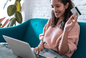 A woman sits on the couch with a laptop in her lap and holding a credit card.