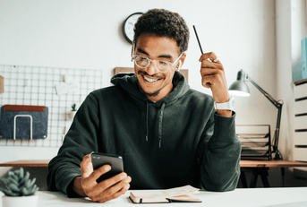 A man sits at his office, smiling down at his phone. 