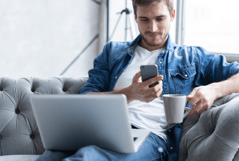 A man sits on the couch and uses his phone and laptop.