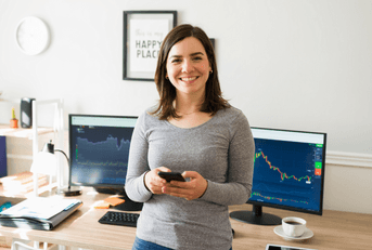 A woman holds her phone while smiling in front of computers.