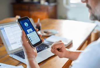 Man checking online bank account balance on smartphone while working on laptop at home