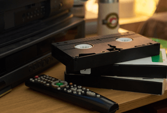 Close-up of several vintage VHS tapes stacked near remote control on wooden table with an old TV