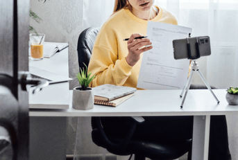A young woman holds up a piece of paper as she speaks in front of her iPhone on a tripod. 