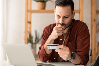 A man appears worried as he holds his chin and looks down at his credit card while sitting in his home office.