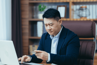 A man stares down at his credit card with his brows furrowed while sitting in his home office.