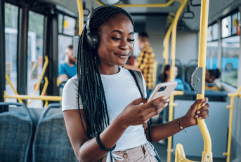A woman wears headphone and smiles while using her smartphone on a bus.