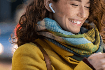 A woman wears wireless earbuds and a scarf while smiling and staring down at her phone.